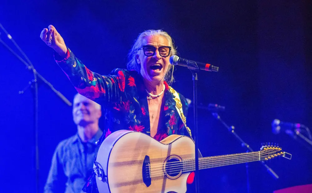 Musician singing and playing a 12-string guitar on stage with blue lighting.