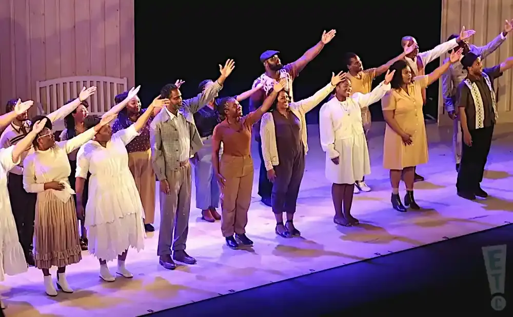 A diverse group of performers stand on stage with arms raised in unison, bathed in stage lighting.