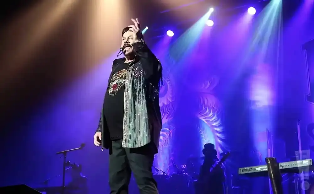 Singer on stage in a black jacket with sparkly fringe and a patterned shirt, performing under purple lights.