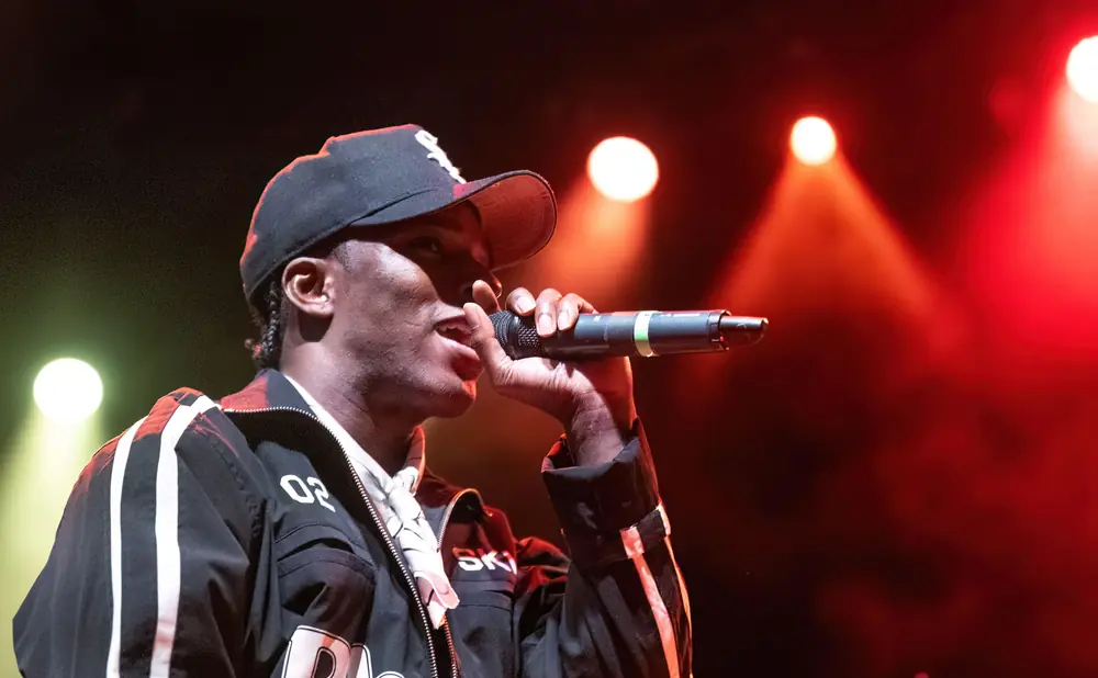 A rapper in a black cap and jacket performs into a microphone under stage lights.