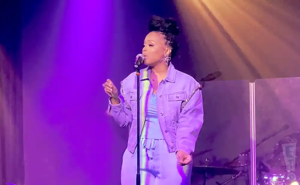 A woman with her hair in a bun sings into a microphone on a stage with purple lighting.