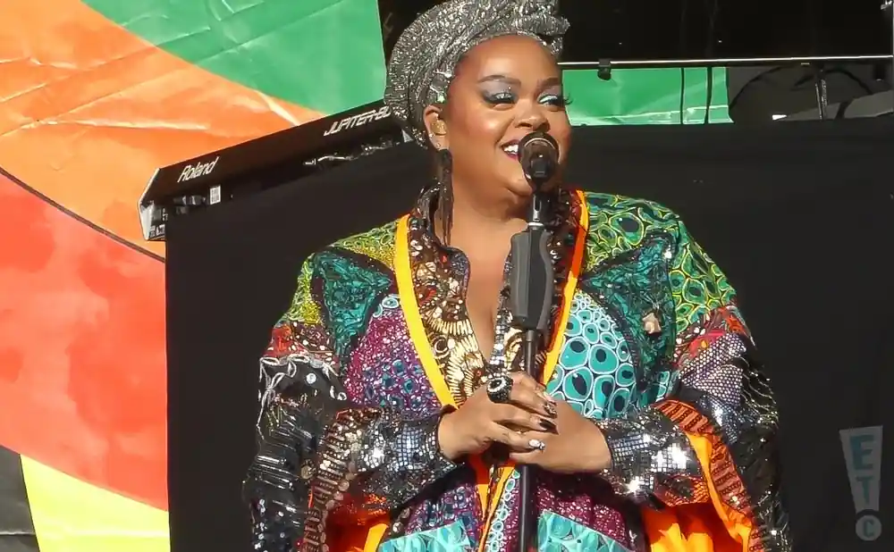 Jill Scott wearing a brightly patterned jacket and silver headwrap sings into a microphone on stage.