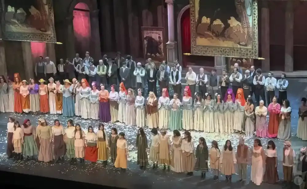 A large cast stands on stage, a mix of men and women in period costumes, bowing to the audience amidst scattered confetti.