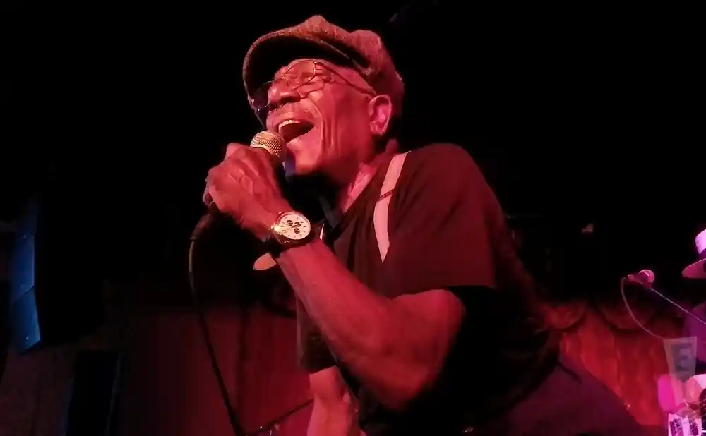 A man in a cap and glasses sings into a microphone, bathed in red light.