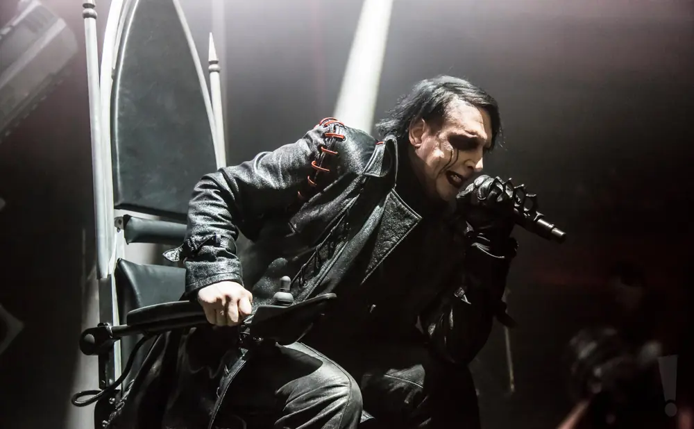 Marilyn Manson sings into a microphone on stage, dressed in black leather and makeup.
