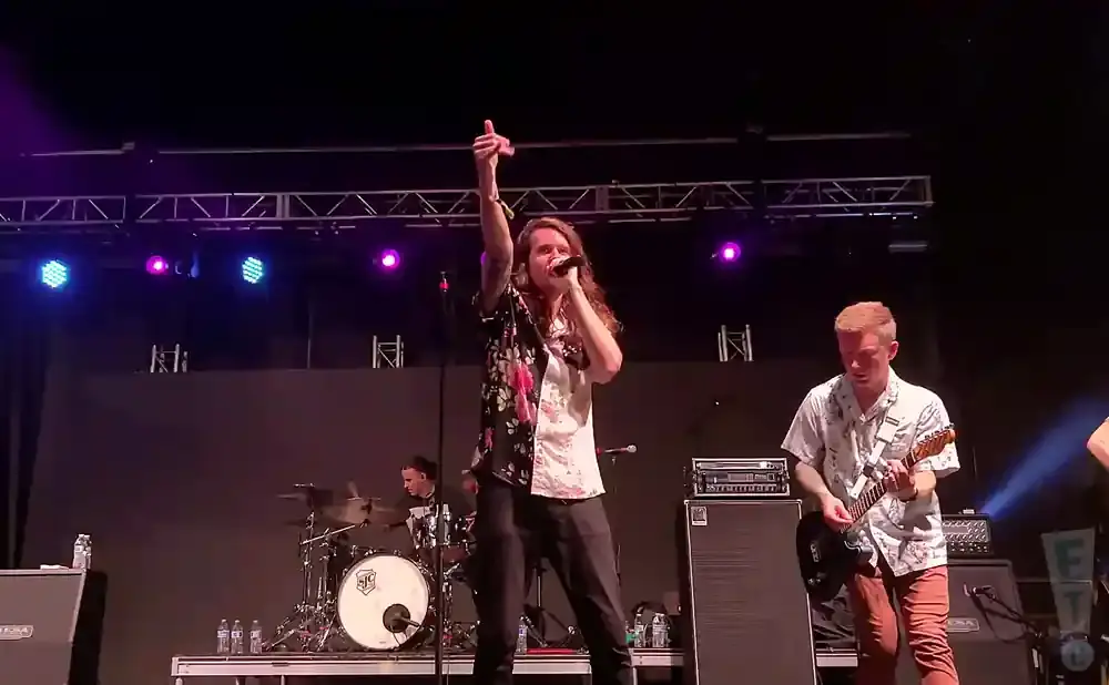 A male singer with long brown hair in a floral shirt points up while singing into a microphone on stage.