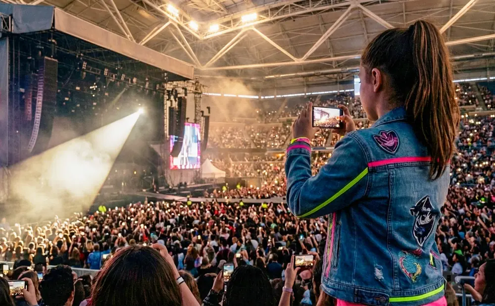 A girl films a concert on her phone, wearing a denim jacket with colorful patches.