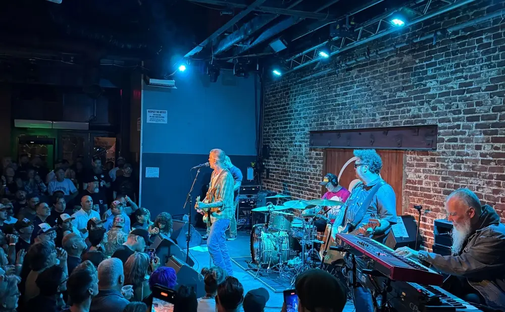 Musicians perform on a dimly lit stage with a brick wall background, in front of a cheering crowd.