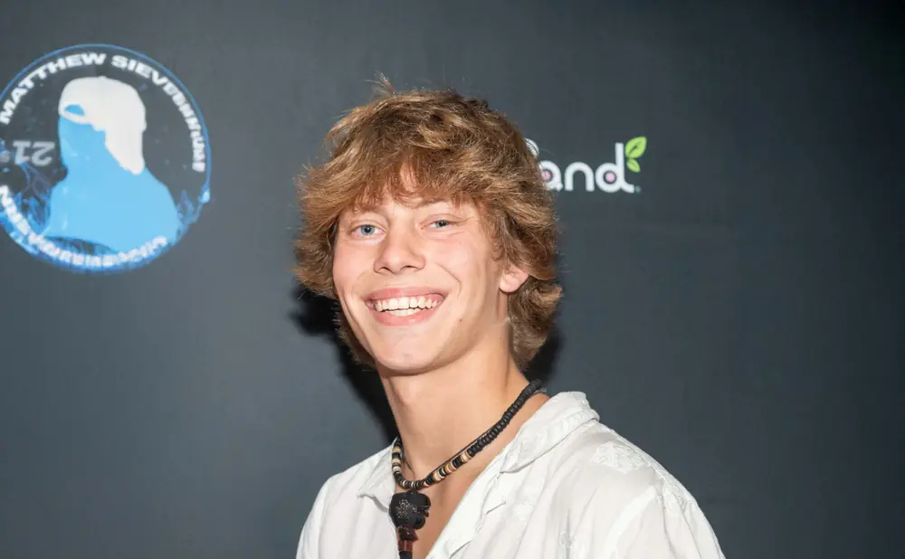 A young man with light brown hair and blue eyes smiles at the camera, wearing a white shirt and a beaded necklace with a pendant.