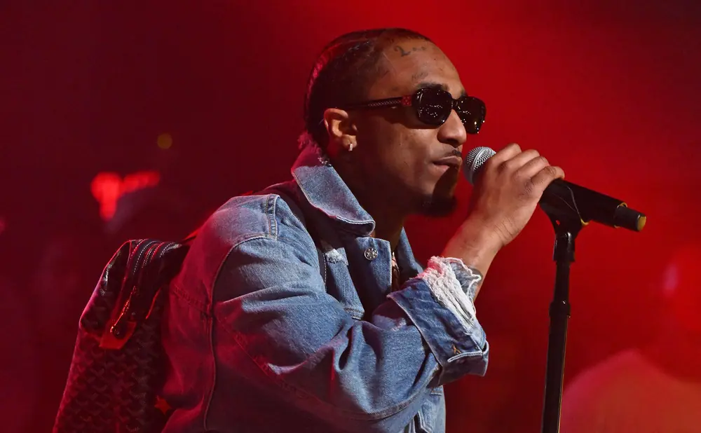 A man with sunglasses and a denim jacket sings into a microphone on a red-lit stage.