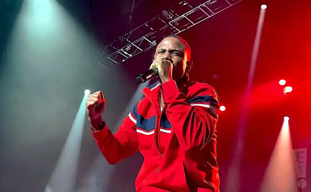 A Black man in a red jacket raps into a microphone on a stage with red and white lights.