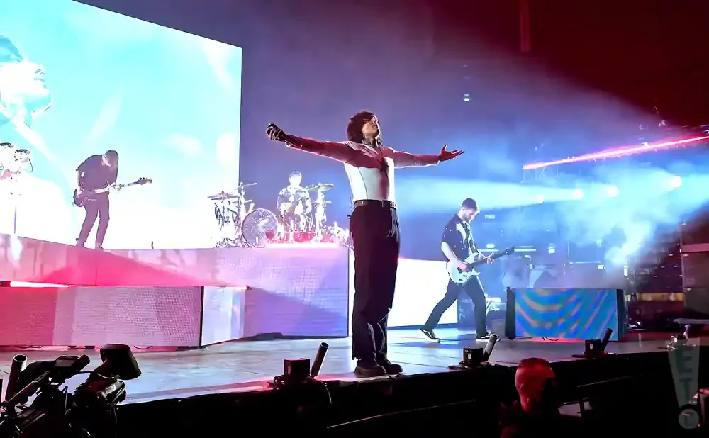 A lead singer stands with arms outstretched on stage with a band playing behind him.