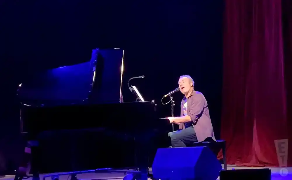 A man sings while playing a grand piano on a stage with a dark background and red curtains.