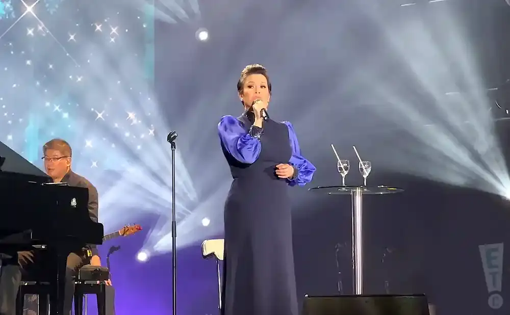 Singer in a dark blue dress with voluminous blue sleeves performs with a microphone on stage, accompanied by a pianist.