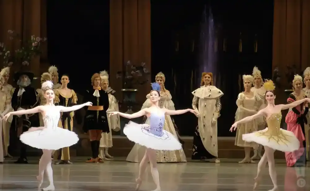 Ballet dancers in colorful tutus on stage with an ensemble of costumed performers.