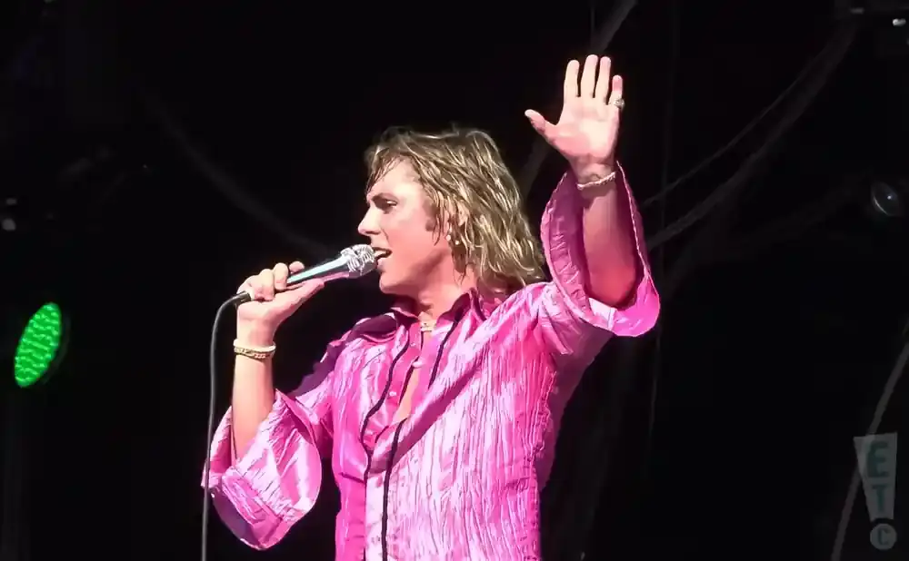 A singer in a pink shirt holds a microphone and raises his hand on stage.
