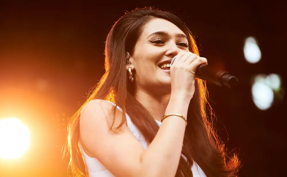 A woman with long brown hair smiles while singing into a microphone on a stage.