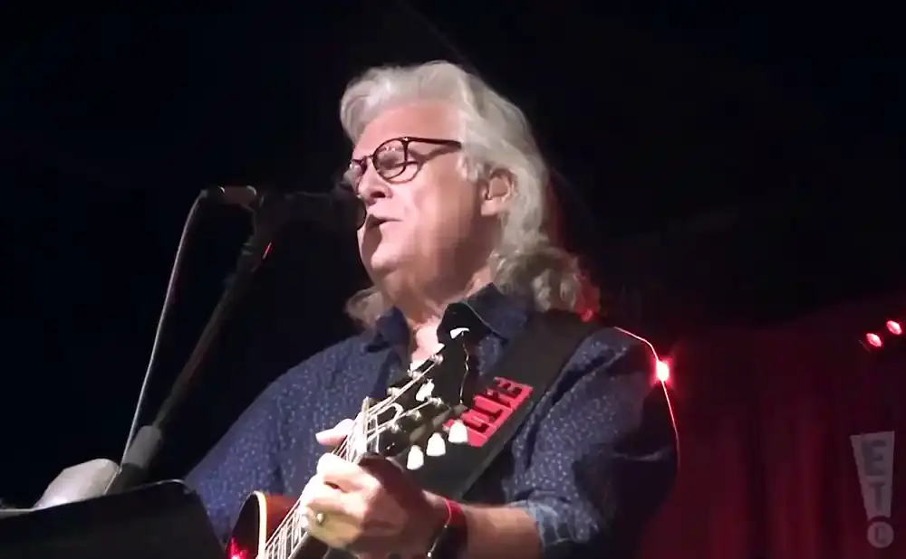 Musician with long gray hair and glasses playing guitar on stage.