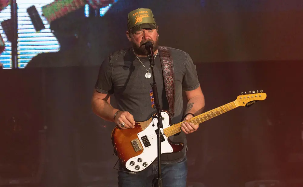 Lee Brice performs on stage, wearing a green hat and playing a guitar.