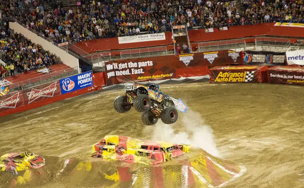 A monster truck leaps over crushed cars in a dirt arena with a large crowd watching.