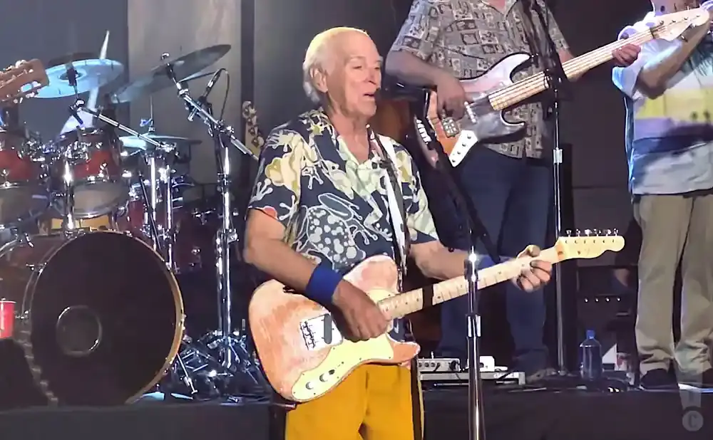 Man in a Hawaiian shirt plays a light-colored electric guitar on stage, with a drum set and other musicians in the background.