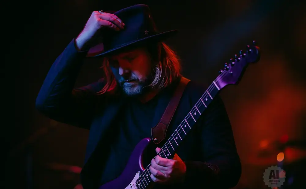 A guitarist in a black hat plays a purple electric guitar under colorful stage lights.