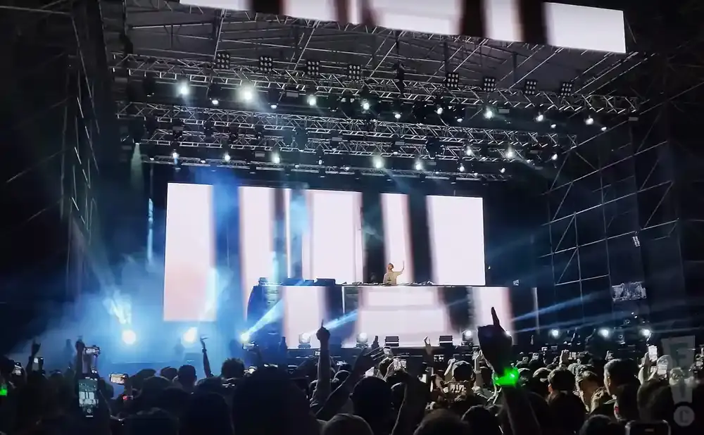A DJ performs on stage at a concert with a large crowd in the foreground.