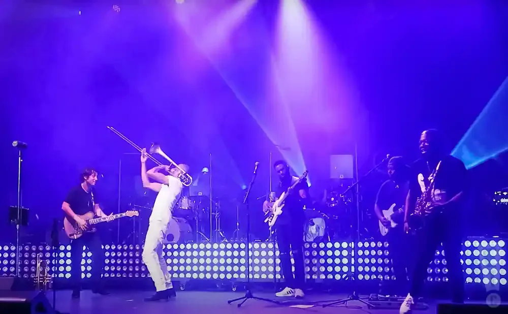 A band performs on stage with purple lights, featuring a trombonist in white, guitarists, a saxophonist, and a drummer.
