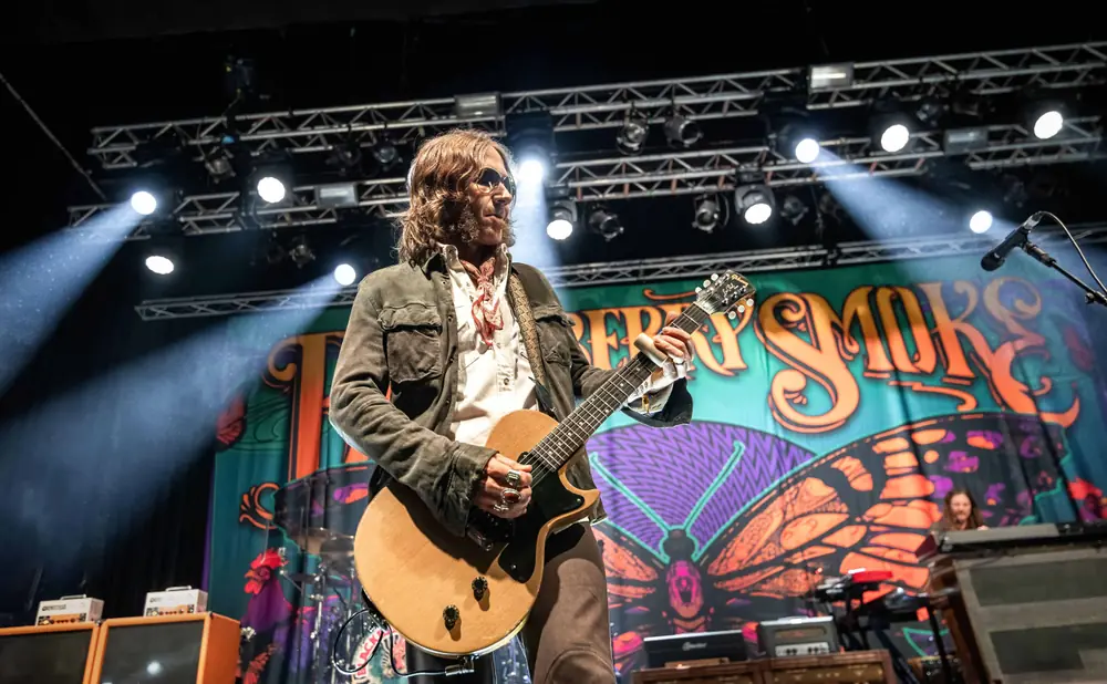 A guitarist on stage, illuminated by spotlights, plays a wooden electric guitar in front of a colorful butterfly backdrop.