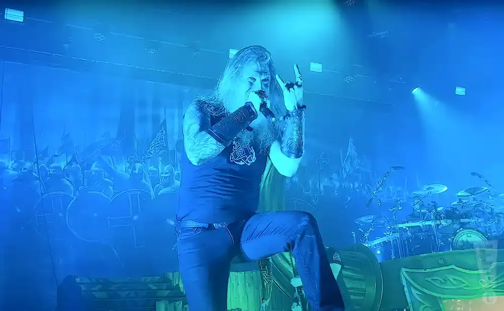 Man with long hair and beard, wearing dark clothing and holding a microphone, performs on stage with blue lighting and a Viking-themed backdrop.