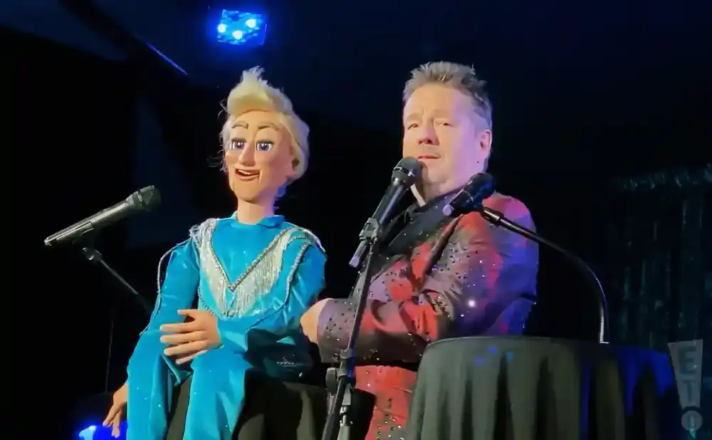 A ventriloquist performs with a blonde puppet wearing a blue dress, both in front of microphones on a stage.