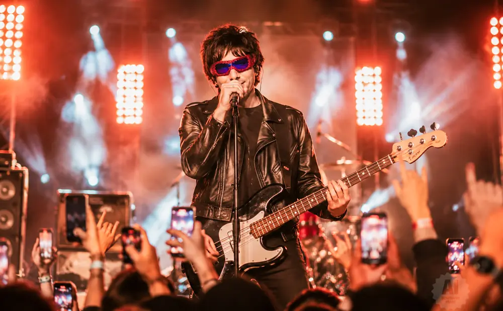 A rock singer in a leather jacket and sunglasses performs on stage, playing a bass guitar in front of a crowd with phones raised.