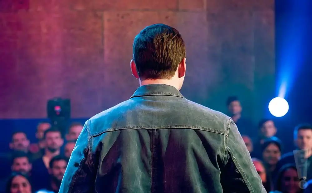 Man in denim jacket facing away from camera, on stage with audience in background.
