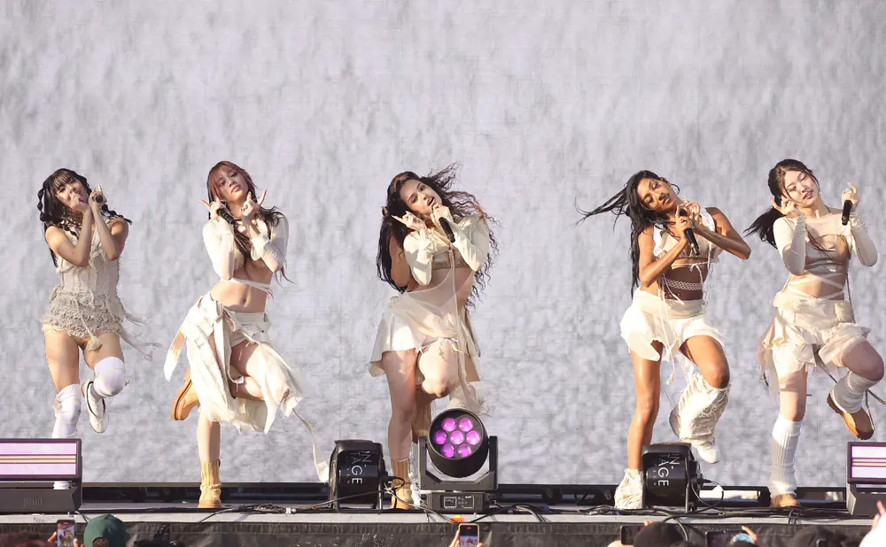 Five women in light-colored outfits dance on a stage in front of a large, bright screen.