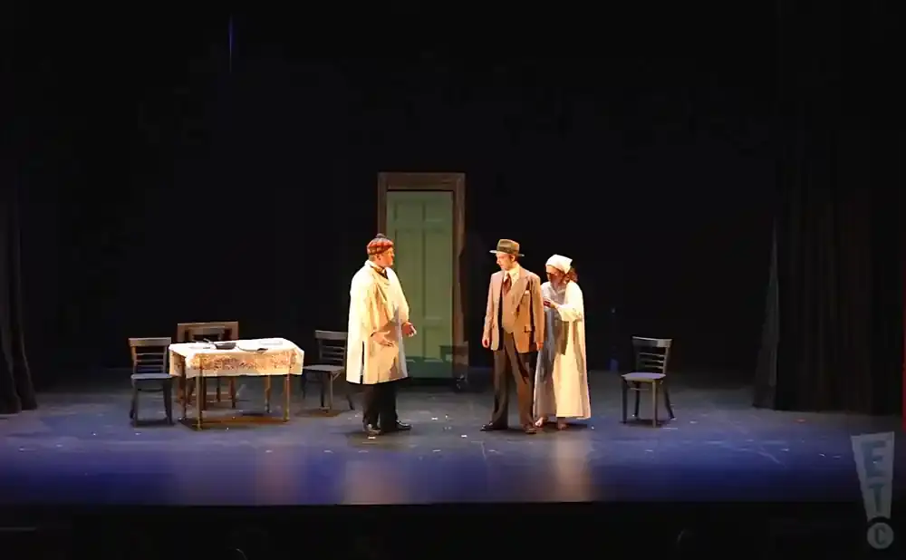 Stage actors in period clothing interact on a set with a door, table, and chairs.