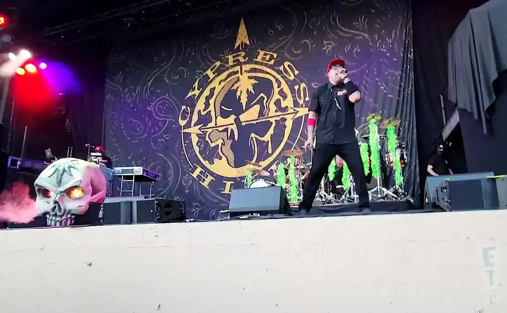 A Cypress Hill band member performs on stage in front of a large banner, with a skull prop emitting smoke.