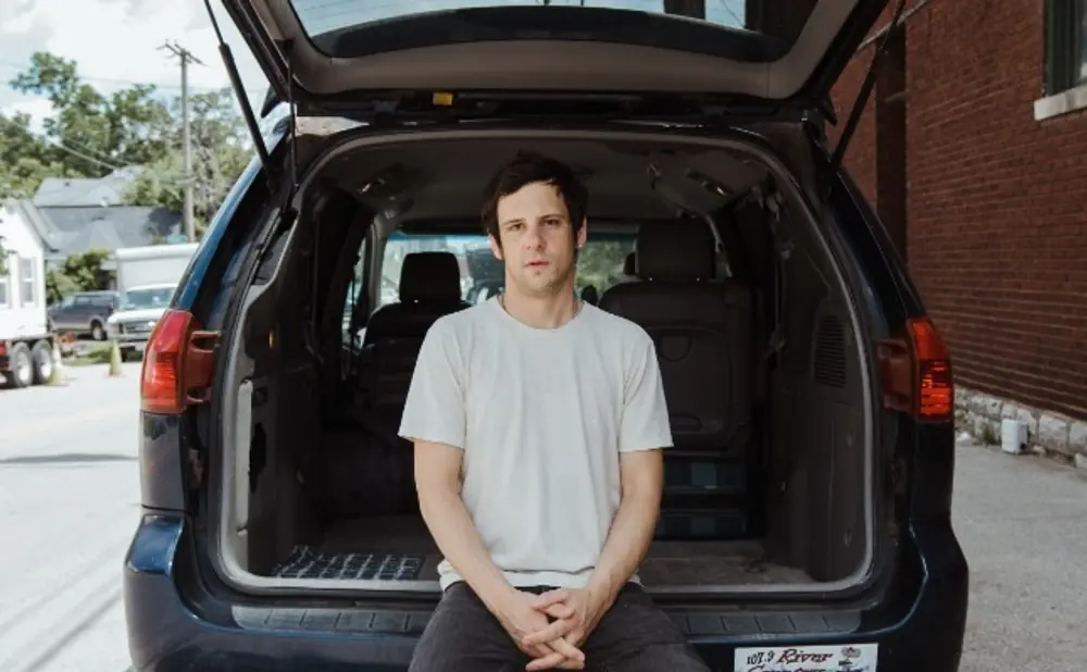 Man in white t-shirt sits in the open trunk of a dark minivan, looking at the camera.