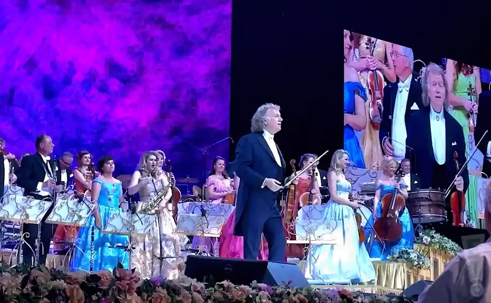 A conductor leads a classical orchestra on stage with a large screen displaying a close-up of the conductor.