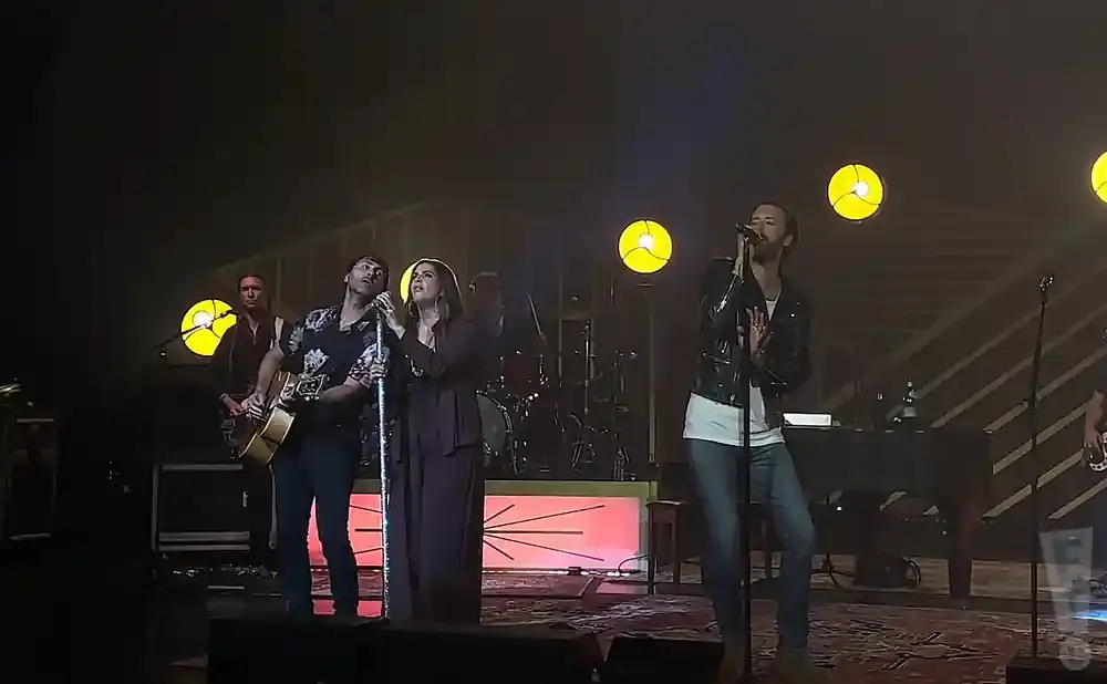 A band performs on stage, with singers at microphones and a guitarist.