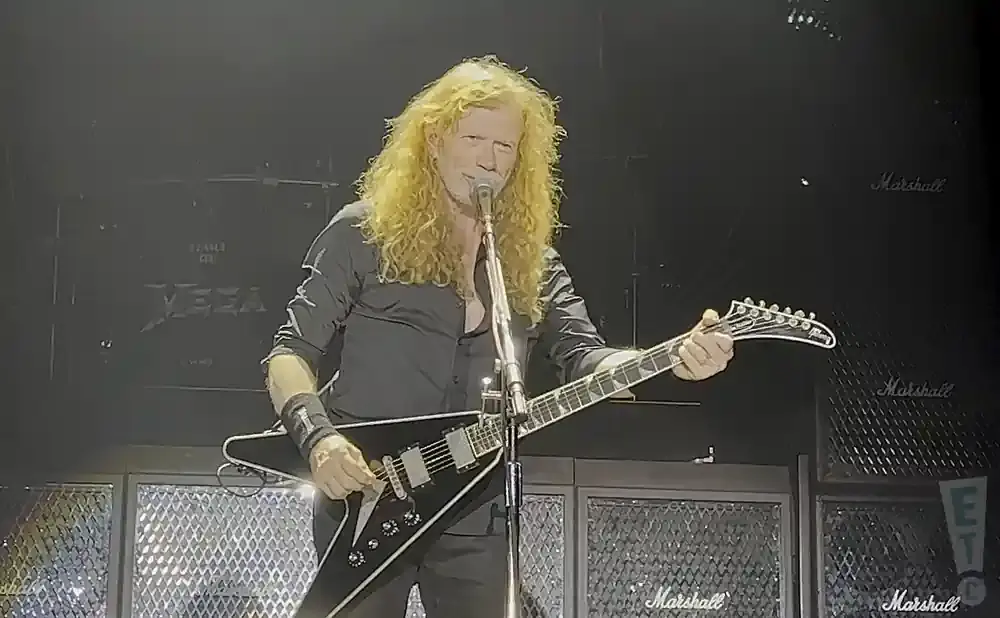 Dave Mustaine of Megadeth plays a black V-shaped guitar on stage with Marshall amps behind him.