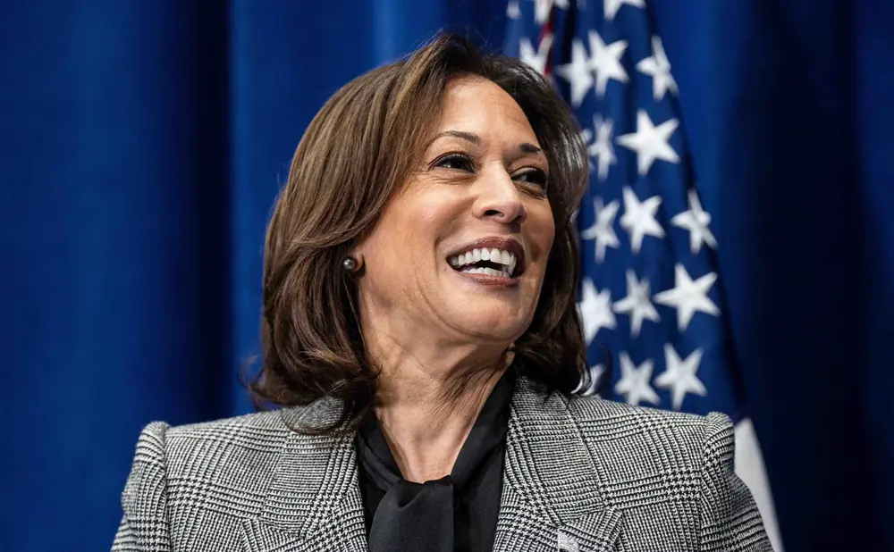 Kamala Harris smiles in a grey plaid jacket and black shirt in front of an American flag.