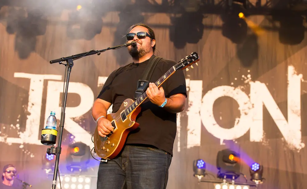 A guitarist with sunglasses and a beard plays an electric guitar on stage.
