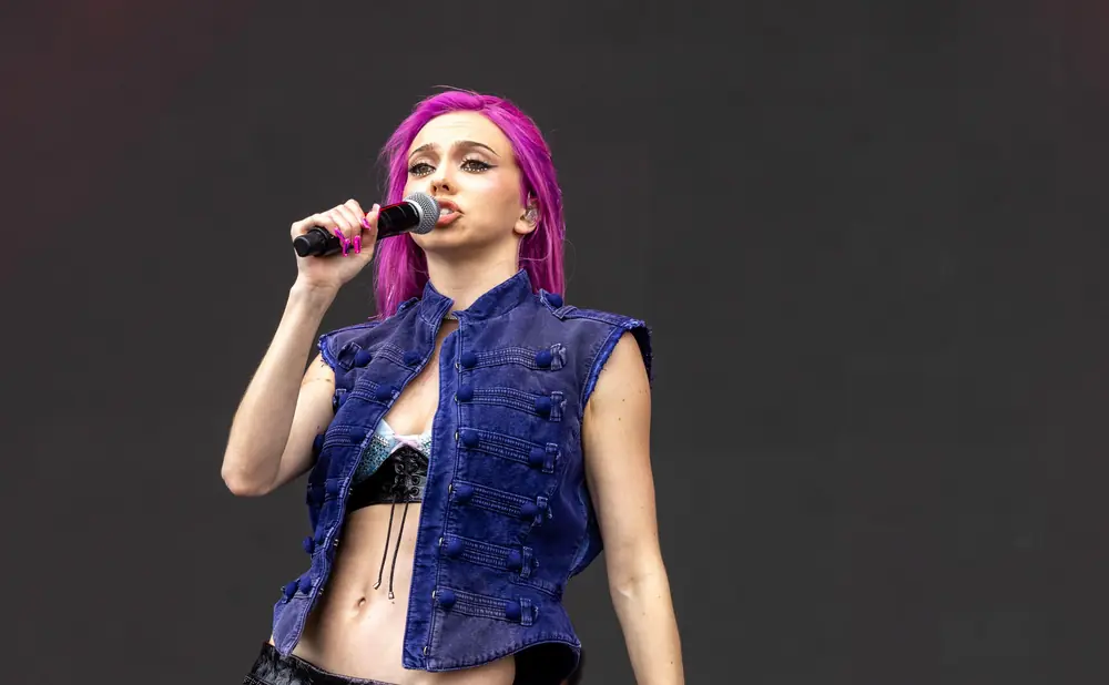 A woman with bright pink hair sings into a microphone while wearing a blue denim vest over a sparkly top and black skirt.