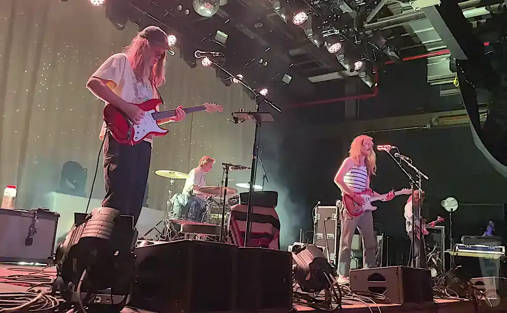 A band plays on stage, with a guitarist in the foreground and drums and another guitarist in the background.