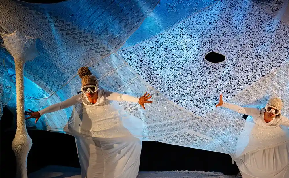 Two performers in white, wearing goggles and beanies, with arms outstretched on a stage draped with white lace fabric.