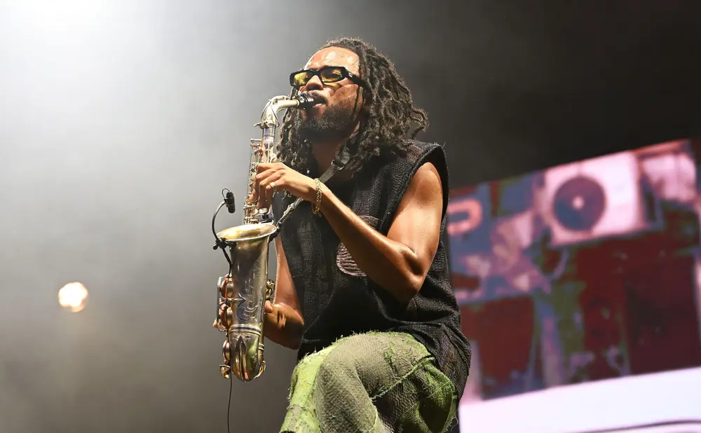 A man with dreadlocks plays a saxophone onstage, wearing sunglasses and green pants.