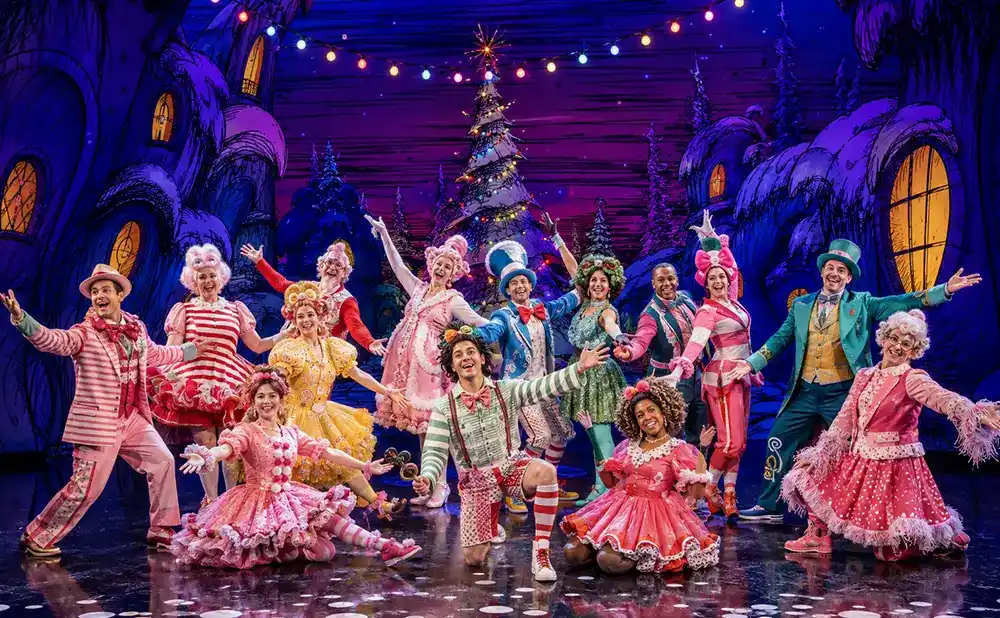 A group of performers in whimsical, colorful costumes on a stage with a fantastical, cartoonish backdrop.