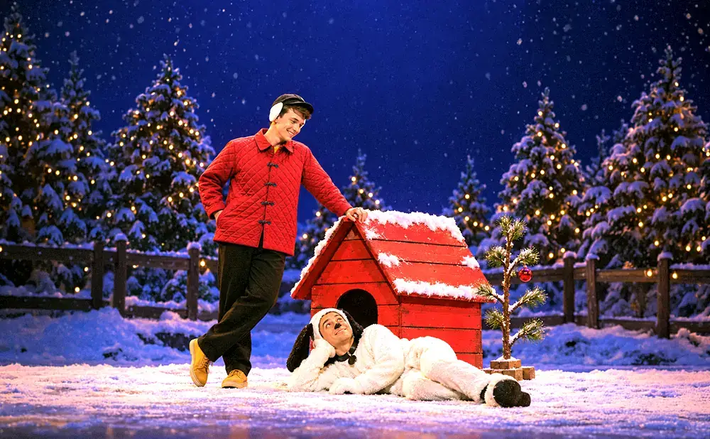 Two actors on a snowy stage: one in a red jacket, the other in a Snoopy costume.