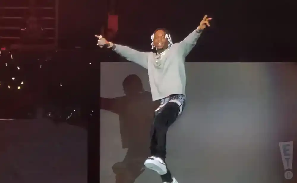 A Black man with white braided hair, wearing a gray sweatshirt and black pants, dances on stage with his arms raised.