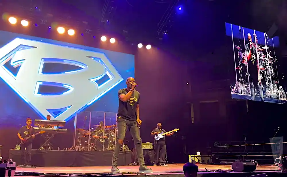 A Black male singer in a Superman-style shirt performs on stage with a band, with a large 